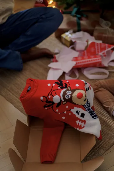 Pull de Noël rouge à tête de renne au nez en pompon, sorti de son carton d'expédition parmi des papiers cadeaux déchirés
