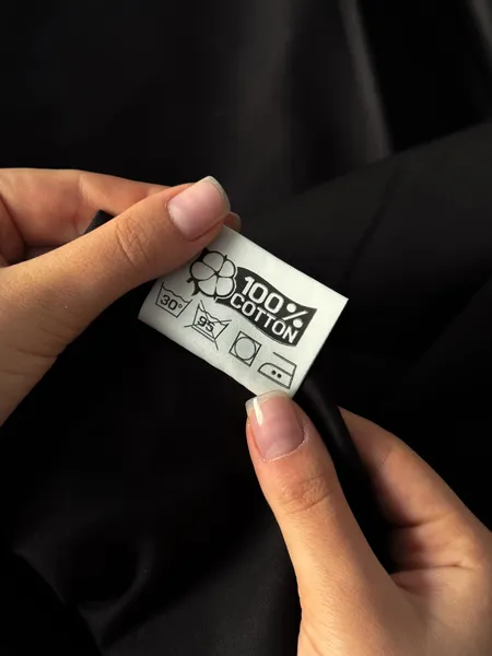 Étiquette « 100 % Cotton » tenue entre deux doigts : lavage à 30 °C, lavage à 95 °C barré, sèche-linge et repassage à deux points