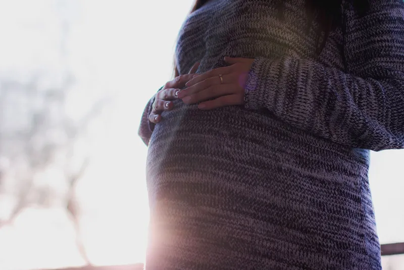 Femme enceinte en pull chiné gris et violet, les mains posées sur le ventre, en contre-jour