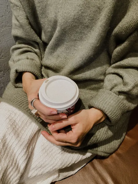 Mains serrées autour d'un gobelet de café, dans les manches longues et amples d'un pull en maille vert sauge