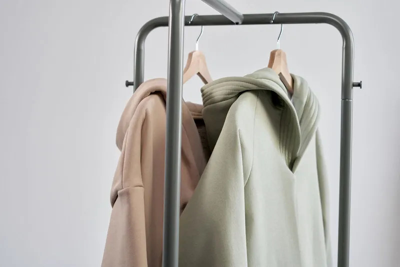 Deux sweats à capuche, beige et vert amande, sur cintres en bois à un portant métallique