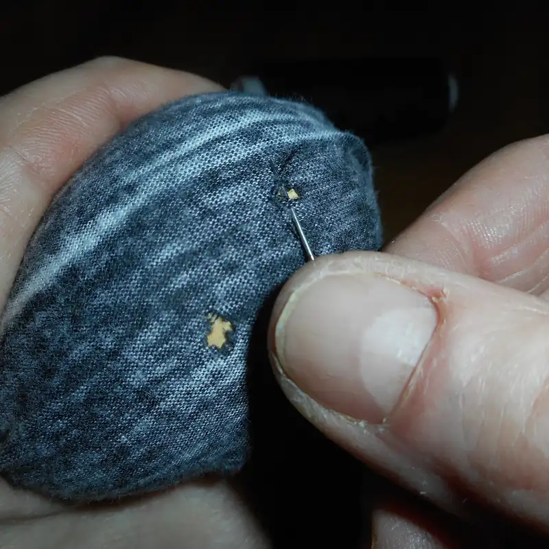 Deux mains tendent un tricot gris chiné sur un champignon à repriser, une aiguille reprend un petit trou